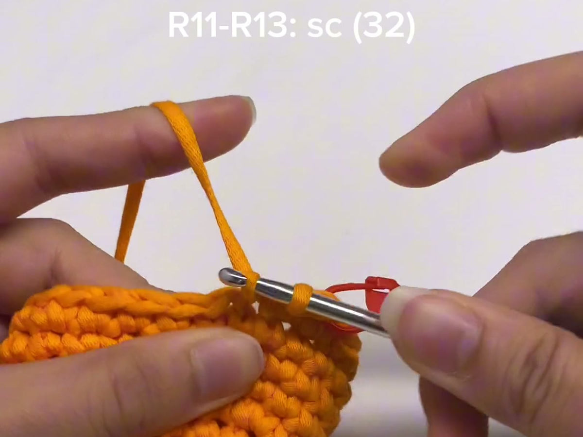 Orange yarn feeding over the fingers into a round of single crochet, with the pattern line R11-R13: sc (32) shown above
